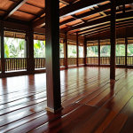 A beautiful traditional Malay wooden house interior, focusing on the elevated, polished dark hardwood floor. Natural sunlight streams through the windows, highlighting the intricate wood grain and casting soft shadows. Below the floor, a visible airy space suggests optimal natural ventilation, creating a cool and tranquil atmosphere within the tropical setting. The scene should convey a sense of timeless comfort and ingenious climate adaptation, without any modern cooling systems.